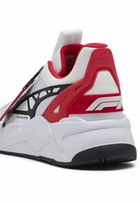 Biała sportowa tenisówka z czerwonymi i czarnymi akcentami. Posiada fakturowaną cholewkę, wyściełany zapiętek oraz widoczną, zakrzywioną podeszwę z rowkami.