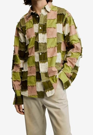 Homme portant une chemise à manches longues boutonnée avec un motif patchwork en vert, rose, blanc et marron, associée à un pantalon beige clair.