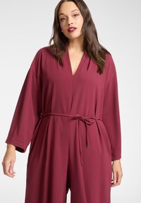 Bordowy kombinezon z głębokim dekoltem w kształcie litery V, długimi rękawami i paskiem w talii. Miękki materiał o gładkiej fakturze oraz boczne kieszenie.