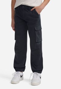 Melnas cargo bikses ar elastīgām piedurknēm, sānu kabatām un pogu aizdari, izgatavotas no izturīga auduma, saskaņotas ar baltām sporta apaviem.