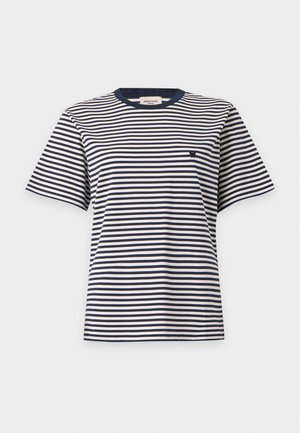 Femme debout portant un t-shirt à manches courtes rayé bleu marine et blanc, un jean bleu et des ballerines noires pointues, sur un fond clair uni.