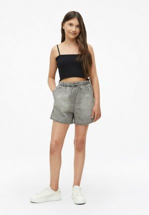 Jeune fille aux longs cheveux bruns portant un top court noir, un short gris taille haute et des baskets blanches, debout avec une main dans la poche.
