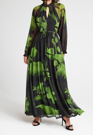 Femme portant une robe maxi noire transparente avec de grands imprimés floraux verts, des manches longues, un décolleté clé et des sandales noires à talons hauts.