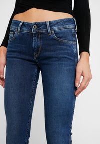 Mörkblå denimjeans med slim fit, femfickorsdesign och kontrasterande orange sömnadsdetaljer. Midjehöjd med knäppning.