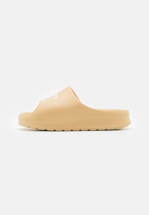 Chancla deslizante beige con parte superior sintética suave y suela texturizada. Presenta un diseño minimalista con forma contorneada y punta abierta.