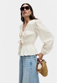 Hvid tekstureret bluse med V-hals, rynket krop og bindedetalje; kombineret med lyseblå bukser med brede ben og en flettet taske.