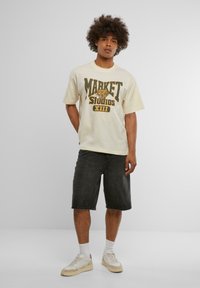 Krémové bavlněné tričko s černou a žlutou grafikou "MARKET STUDIOS", doplněné tmavými džínovými šortkami a světlými teniskami s krémovými akcenty.