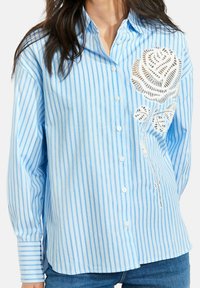 Kobieta ubrana w jasnoniebieską i białą koszulę w paski zapinaną na guziki z dużym białym koronkowym wzorem róży po lewej stronie, w połączeniu z niebieskimi dżinsami.