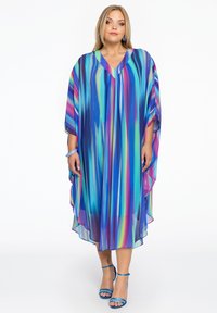 Kaftankleid in farbverlaufenden Streifen von Blau, Grün, Pink und Lila. V-Ausschnitt-Design, fließender Stoff und weite Ärmel. Blaue Riemchensandalen.