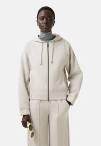 Beige zip-hoodie med snor i huen, der har forlommer, ribbede manchetter og en blød tekstur, parret med matchende brede bukser.