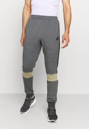 Mann trägt graue Adidas Jogginghosen mit beigen Knie-Patches und schwarzen Adidas Sneakers, stehend vor einem einfarbigen hellen Hintergrund.