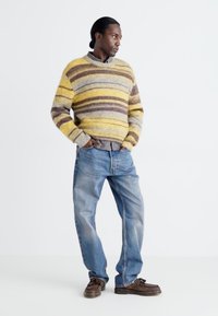 Suéter a rayas multicolores en amarillo, marrón y gris, combinado con jeans de mezclilla azules y zapatos de cuero marrón. Corte casual y tejido texturizado.