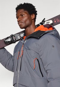 Veste isolante grise avec une capuche orange, dotée de zips noirs et de cordons réglables. Le modèle porte des skis sur l'épaule.
