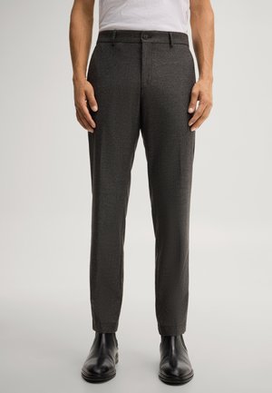 Man draagt een slim-fit donkergrijze nette broek en zwarte leren enkellaarzen, staand tegen een effen lichte achtergrond.