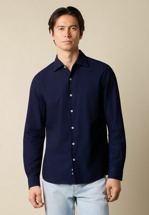 Młody mężczyzna stojący, ubrany w ciemnoniebieską koszulę na guziki i jasnoniebieskie dżinsy na tle jednolitego beżowego tła.