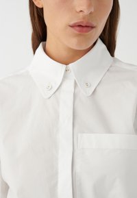 Camicia bianca in cotone con colletto classico, bottoni frontali e taschino sul petto. Il tessuto ha una consistenza liscia e croccante.