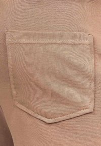 Primo piano di un tessuto beige che presenta una tasca a toppa con un bordo cucito dritto e una texture liscia. Il tessuto ha una leggera texture.