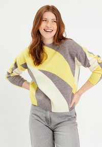 Pull en tricot gris, jaune et blanc avec des rayures diagonales audacieuses, des poignets et un ourlet côtelés, offrant une coupe décontractée et une texture douce.