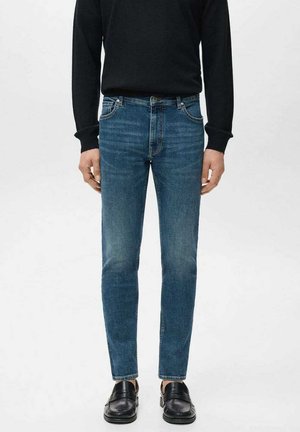 Vaqueros ajustados de mezclilla azul oscuro, con bolsillos delanteros, detalles de desgaste ligero y un corte slim, combinados con zapatos de vestir negros y brillantes.