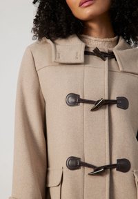 Manteau en mélange de laine beige avec un design à double boutonnage, fermetures à bascule marron, poches latérales et un col haut. Texture lisse.