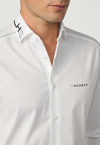 Witte overhemd met een slanke pasvorm, een button-down kraag, voorzien van zwarte branding "HACKETT" en een contrasterend kraagontwerp. Gladde stofstructuur.