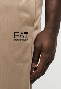 Μπεζ ελαφριά παντελόνια από ύφασμα με υφή. Διαθέτουν μαύρη επιγραφή "EA7 EMPORIO ARMANI" στη πλευρά. Κοντινό πλάνο ενός χεριού που ακουμπάει στο ύφασμα.