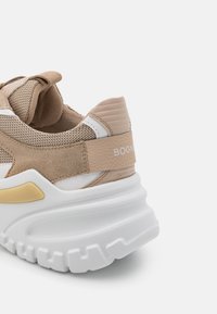 Sneaker beige con tomaia in rete e suede, intersuola bianca con scanalature testurizzate e un sottile accento dorato vicino alla suola.