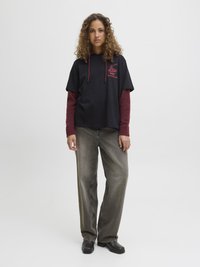 Haut à manches courtes noir avec logo rouge, superposé sur une chemise à manches longues bordeaux, associé à un jean en denim gris ample et des chaussures noires.