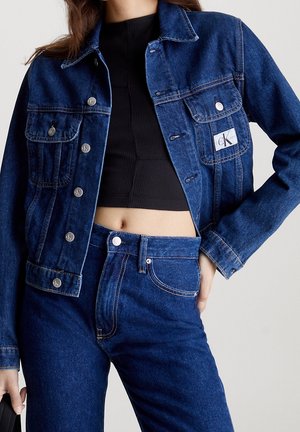 Femme portant une veste en jean bleu foncé et un jean taille haute avec des boutons argentés, associé à un top court noir côtelé.