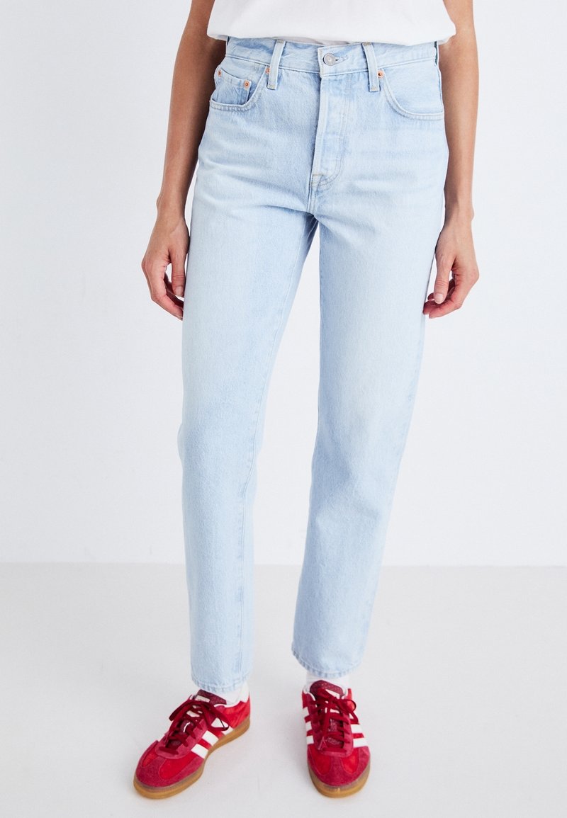 Personne portant un jean taille haute bleu clair, un haut blanc, des chaussettes blanches et des baskets rouges à rayures blanches, debout sur un fond uni clair.