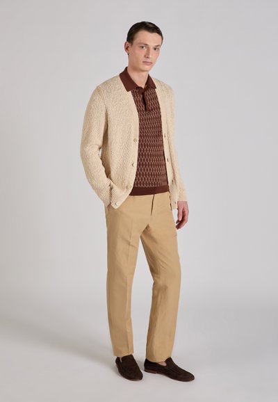 Beige gestrickte Strickjacke mit vorderen Knöpfen, darüber eine braun gemusterte Poloshirt. Kombiniert mit hellen khakifarbenen Hosen und dunklen Loafers.