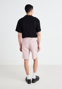Chemise noire à manches courtes associée à un short rose clair, avec une texture lisse et deux poches arrière. Chaussures noires et chaussettes blanches visibles.