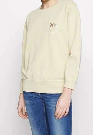 Persoon draagt een crèmekleurige sweater met een metalen vlindelogo op de borst en blauwvervaagde skinny jeans, staand tegen een effen achtergrond.