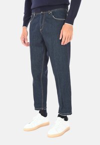 Pantaloni in denim scuro con vestibilità rilassata, dotati di cuciture arancioni a contrasto, cinque tasche e orlo arrotolato, abbinati a sneakers bianche.