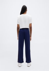 Άτομο με μακριά σκούρα μαλλιά στέκεται με την πλάτη γυρισμένη, φορώντας ένα κοντό λευκό μπλουζάκι, μπλε σκούρο παντελόνι φαρδύ με ρίγες στο πλάι και λευκά αθλητικά παπούτσια.