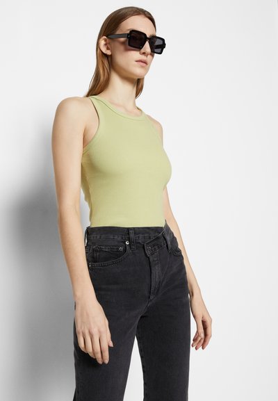 Ribbed tank top in light green, paired with high-waisted black jeans. Model wearing large, rectangular black sunglasses, standing against a white background.