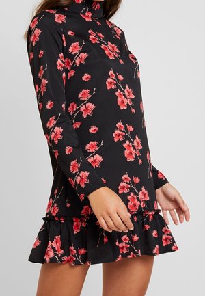 Femme portant une robe noire à manches longues avec un imprimé floral rouge et un ourlet à volants, debout devant un fond gris uni.