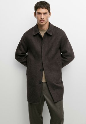 Giovane uomo con capelli corti e scuri che indossa un cappotto di lana marrone scuro sopra un maglione marrone chiaro e pantaloni oliva, in piedi con le mani dietro la schiena.