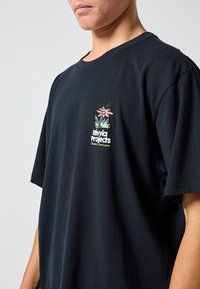 T-shirt de algodão preta com um desenho floral em rosa e verde na área do peito esquerdo, com o texto "Rivvia Projects" abaixo do gráfico.