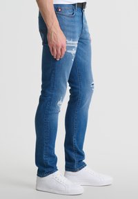 Lieknos mėlynos džinsinės kelnės su dėvėjimosi detalėmis, turinčios klasikinį penkių kišenių dizainą ir kontrastingą siuvinėjimą, derinamos su baltomis sportinėmis bateliais.