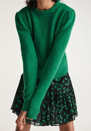 Mujer con un jersey verde difuso de manga larga combinado con una falda negra con un patrón floral verde, brazos cruzados delante.