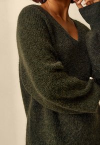 Maglione verde, oversize, con scollo a V profondo. Realizzato in un tessuto morbido e peloso. Presenta maniche lunghe e ampie e una vestibilità rilassata.