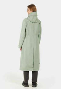 Abrigo largo y ligero de color verde claro, impermeable, con capucha, cintura ajustada y abertura trasera. Presenta un acento en negro del logo cerca del cuello y bolsillos laterales.