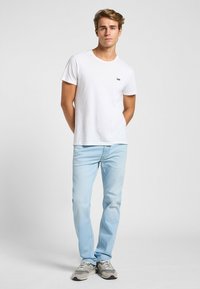 Camiseta blanca de algodón con cuello redondo y un pequeño logo negro de "Lee", combinada con jeans de mezclilla azul claro y zapatillas gris.