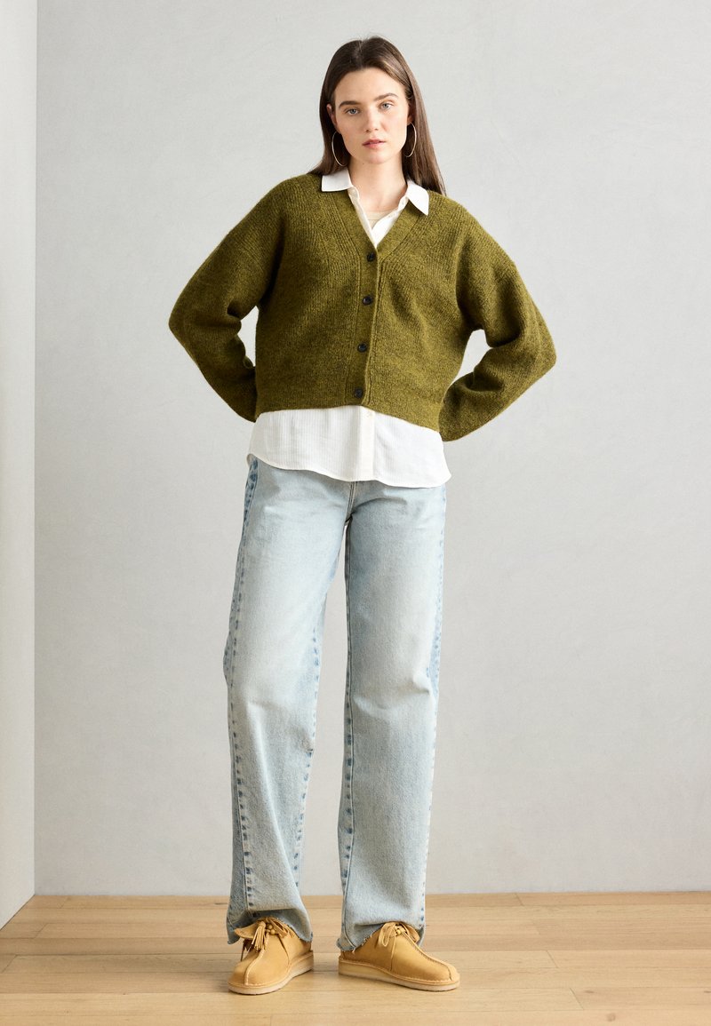 Olijfgroen gebreid vest met een V-hals, knopen aan de voorkant en verlaagde schouders, gecombineerd met een wit shirt en lichtblauwe rechtvallende jeans.