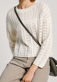 Strickpullover in Creme mit einem strukturierten Muster, ausgestattet mit Rundhalsausschnitt und langen Ärmeln. Kombiniert mit beigen Hosen und einer kleinen grauen Tasche.