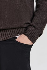 Dunkelbrauner, gestrickter Pullover mit gerippten Bündchen, kombiniert mit schwarzen, eng anliegenden Hosen. Die Hand ruht in der Tasche und zeigt die Stoffstruktur und das Lagenstyling.