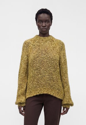 Femme portant un pull en tricot ample jaune moutarde à manches longues et un pantalon marron foncé, debout devant un fond clair uni.