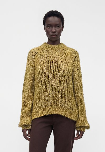 Femme portant un pull en tricot ample jaune moutarde à manches longues et un pantalon marron foncé, debout devant un fond clair uni.