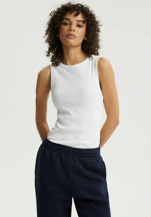 Kobieta o kręconych włosach, ubrana w białą, bezrękawnikową bluzkę i ciemnoniebieskie spodnie, stojąca na jednolitym jasnym tle.